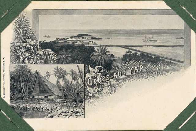 Picture postcard mounted in an album, headed “Gruss aus Yap”: a view over a harbour with steamships and a long straight jetty, framed by drawn orchids and palm fronds, with an inset photograph of a high-gabled thatched house on a stone platform at the water’s edge.