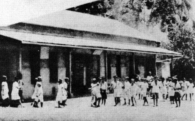 Grainy halftone photograph of a long single-storey school building with a deep veranda roof under tall trees. Groups of children in white clothes walk and stand in the yard in front of it.