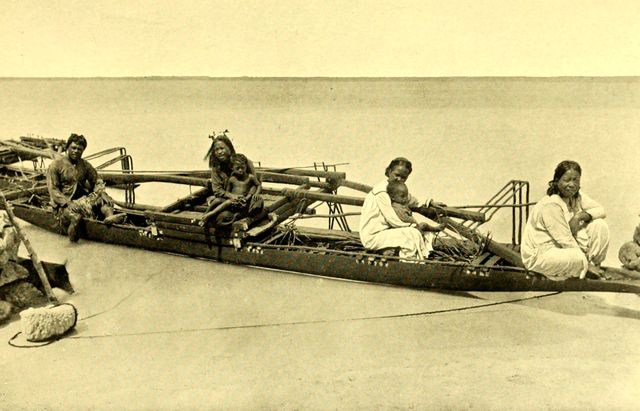 Photograph of a long outrigger canoe drawn up on a sandy beach with the open sea behind. One adult sits near the far end, a girl with a flower in her hair holds a small child on the outrigger platform, and two women in long white dresses sit along the hull, one with a baby in her arms.