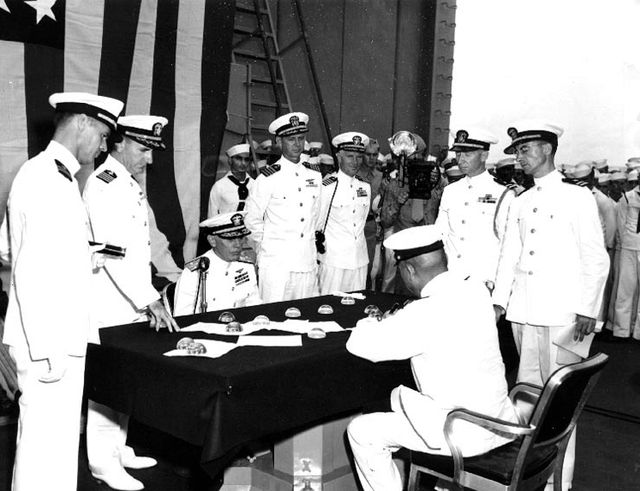 Black-and-white photograph on board a warship. A Japanese officer, seen from behind, signs papers at a cloth-covered table; a US admiral sits facing him, and US Navy officers in white uniforms stand around the table in front of a large US flag.