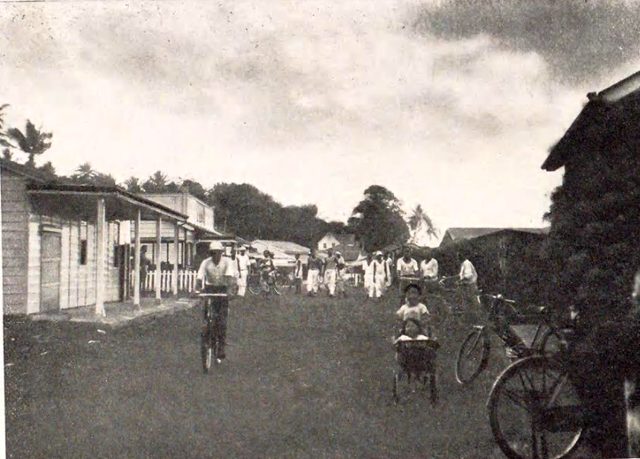 Old black-and-white photograph of a broad dirt street lined with low wooden buildings with verandas; a man rides a bicycle toward the camera, a child sits in a small cart, a bicycle leans in the foreground and people in light clothes stand further down the street under trees.