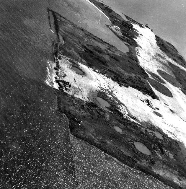 Grainy black-and-white aerial photograph taken at an angle: a flat strip of land along the sea, its surface pale and scarred with dark craters and pools, with a straight seawall and small scattered wreckage along the water's edge. No people are visible.