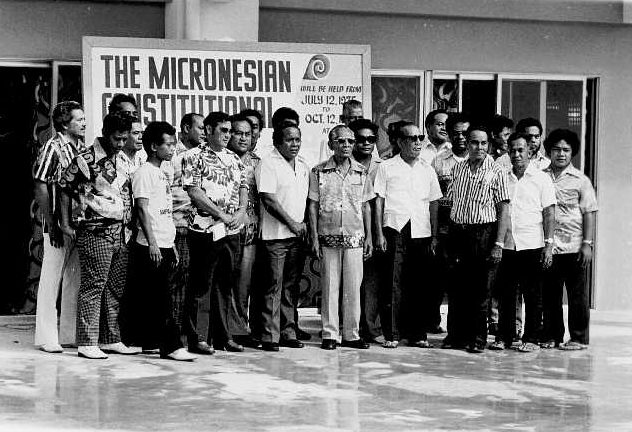 Black-and-white group photograph of about twenty-five men standing in two rows on a shiny floor, many in island-print shirts, in front of a sign reading “The Micronesian Constitutional … will be held from July 12, 1975 to Oct. 12, 1975”.
