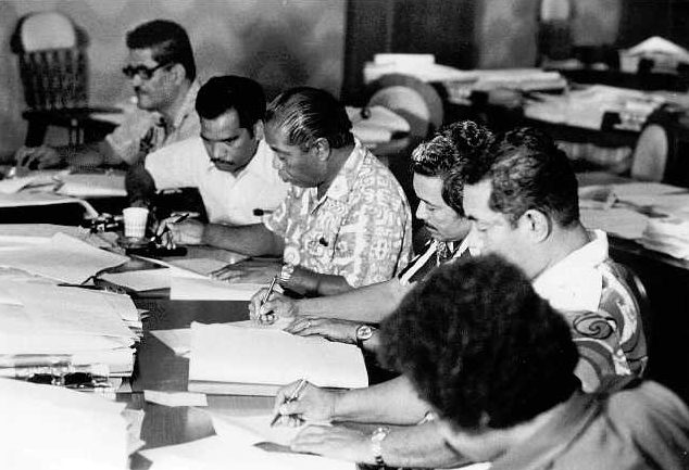 Black-and-white photograph of delegates seated close together along a table covered in papers, several writing with pens; men in island-print shirts and glasses lean over documents, with more desks and stacked papers behind them.