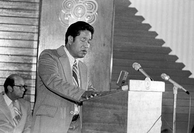 Black-and-white photograph of a man in a light jacket and striped tie reading from papers at a lectern with two microphones; a man in glasses sits to his left, and a carved panel and a zigzag-patterned wall are behind him.