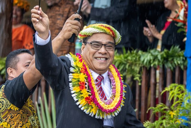 Colour photograph of a smiling man in glasses wearing a woven flower crown and thick layered leis of yellow, red, pink and white flowers over a dark suit, raising a small flag on a stick; people in island dress crowd around him.