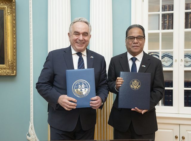 Colour photograph of two men in dark suits standing side by side in a pale blue room with white columns and a gilt-framed painting, each holding up a blue document folder stamped with a gold seal; the man on the left has white hair, the man on the right wears glasses.